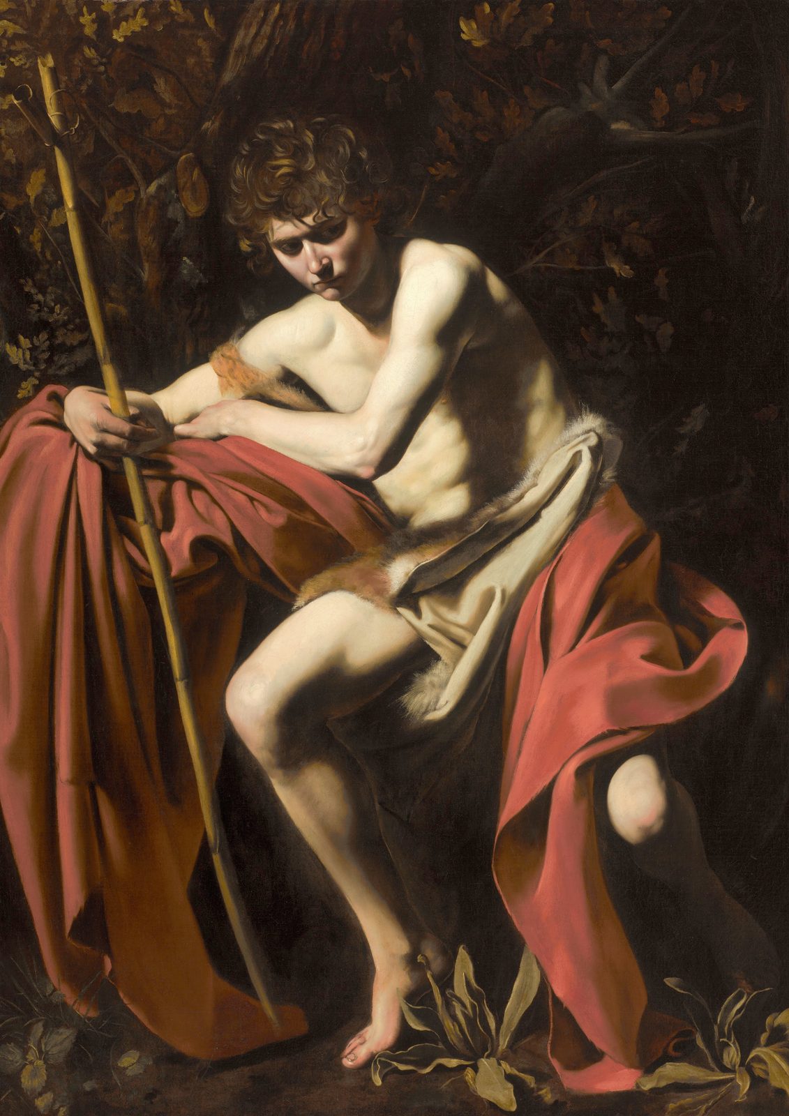 Saint John the Baptist in the Wilderness by Caravaggio, 1604, as a framed print in a black frame on a cream wall. A young man in a scarlet cloak sits in deep shadow holding a thin reed cross, his face turned down in thought, against a dark wilderness.