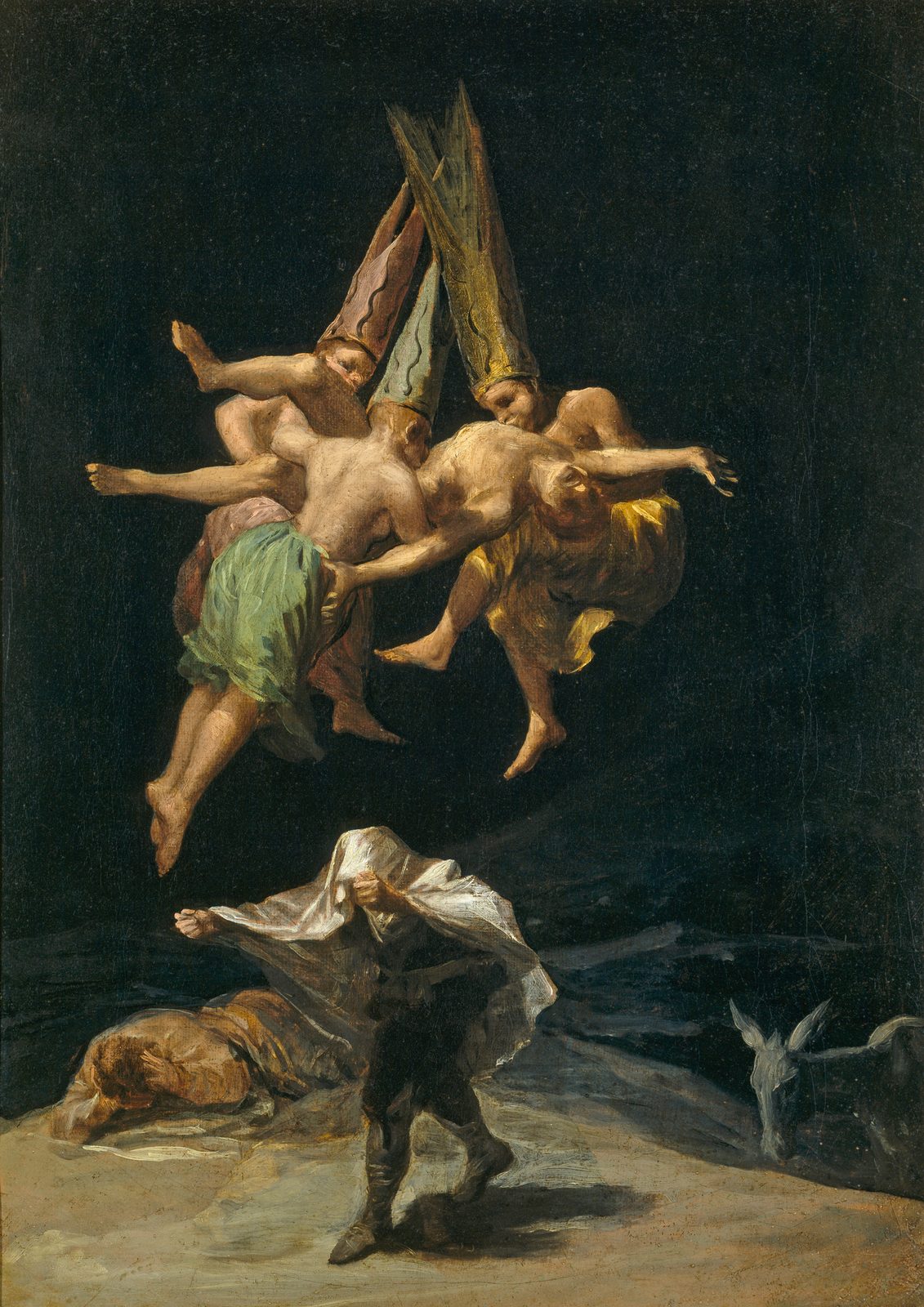 Witches' Flight by Francisco Goya, 1798, as a framed print in a black frame on a cream wall. Three witches in tall pointed hats fly through a black night sky carrying a man, while below one figure hides under a white sheet and another lies on the ground beside a donkey.